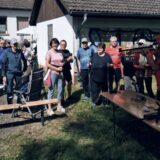 Eine Gruppe überwiegend älterer Menschen steht bei Sonnenschein auf einer Wiese, einige halten Nordic-Walking-Stöcke. Rechts legt ein Mann mit roter Kappe und Weste ein Lasergewehr an. Im Vordergrund stehen Holzbänke und ein Tisch.